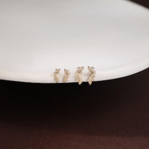 May include: Four gold-toned stud earrings shaped like small fish with detailed fins and scales. The earrings are arranged in a row, featuring two smaller fish on the left and two larger fish on the right, all displaying a metallic finish.
