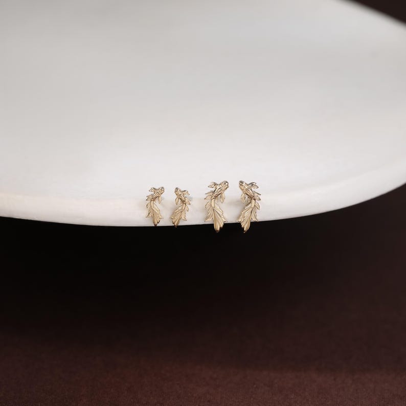 May include: Four gold-toned stud earrings shaped like small fish with detailed fins and scales. The earrings are arranged in a row, featuring two smaller fish on the left and two larger fish on the right, all displaying a metallic finish.