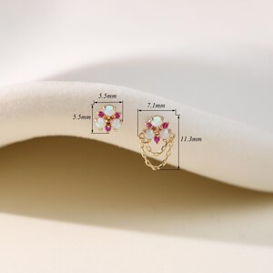 May include: Gold-toned earrings featuring a floral design with white opal and red gemstone accents. One earring measures 5.5mm x 5.5mm, the other 7.1mm x 11.3mm, with a chain detail. The earrings are displayed on a soft, neutral background.