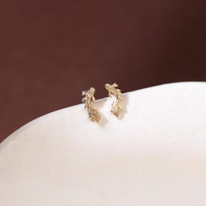 May include: A pair of gold-toned stud earrings shaped like koi fish. Each earring features a detailed, textured design with flowing fins and a curved body, capturing the likeness of a swimming fish in a polished, metallic finish.