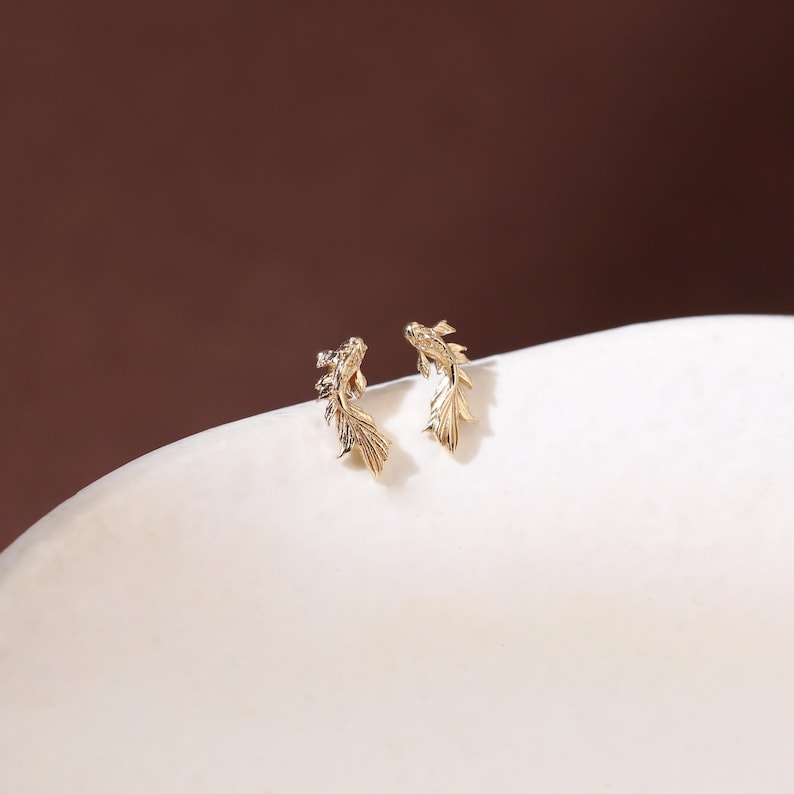 May include: A pair of gold-toned stud earrings shaped like koi fish. Each earring features a detailed, textured design with flowing fins and a curved body, capturing the likeness of a swimming fish in a polished, metallic finish.