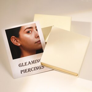 May include: A photograph of a woman's face with multiple ear piercings, displayed on a white card with the words "GLEAMING PIERCINGS" printed in black. The card is next to a closed, cream-colored box.