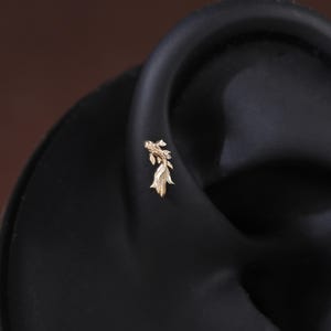 May include: A small gold-toned earring shaped like a koi fish with detailed fins and scales. The earring is positioned on the cartilage of a dark-colored ear model, showcasing the intricate metalwork and the slender, flowing design of the fish.