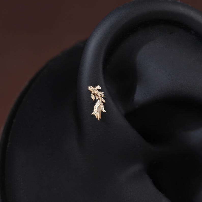 May include: A small gold-toned earring shaped like a koi fish with detailed fins and scales. The earring is positioned on the cartilage of a dark-colored ear model, showcasing the intricate metalwork and the slender, flowing design of the fish.
