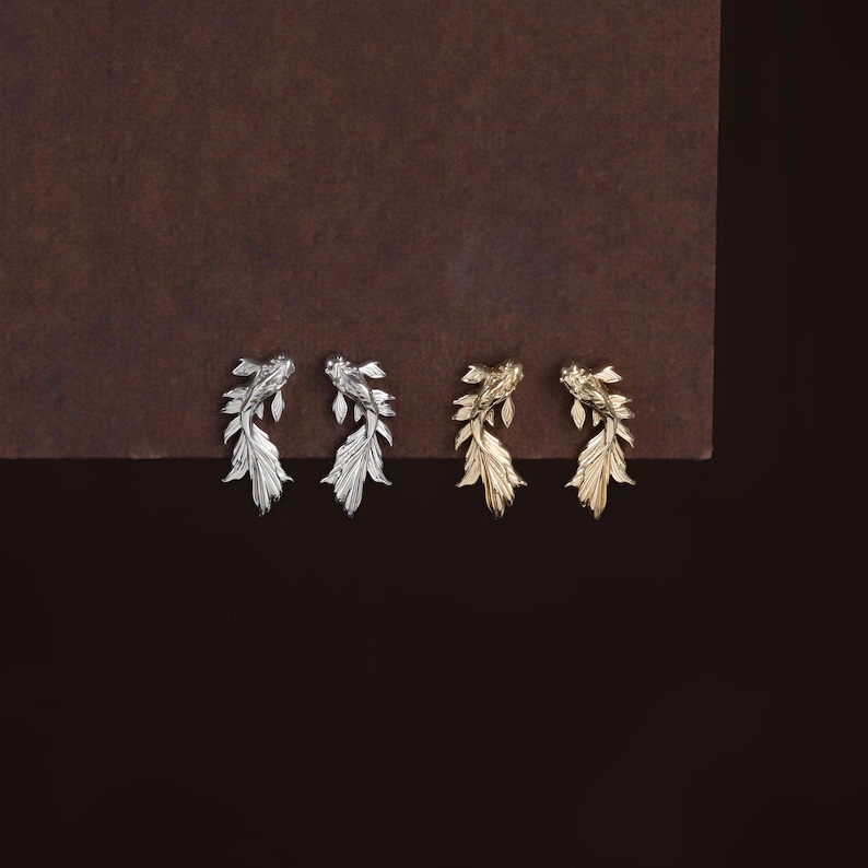 May include: Two pairs of stud earrings shaped like koi fish, one pair in a polished silver tone and one pair in a polished gold tone. Each earring features detailed scales and flowing fins, capturing the swimming motion of the fish in metal.