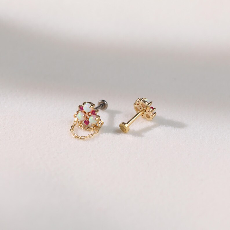 May include: Two gold-colored earrings. One earring features a cluster of white and red gemstones with a small gold chain. The other earring has a similar design, with a gold post and a cluster of gemstones.