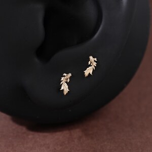 May include: A pair of gold-toned stud earrings shaped like small koi fish. Each earring features detailed fins and a textured body, designed to be worn as a set. The earrings are positioned on a dark, smooth, curved surface that mimics the shape of an ear.