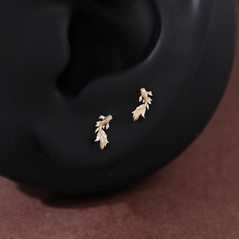 May include: A pair of gold-toned stud earrings shaped like small koi fish. Each earring features detailed fins and a textured body, designed to be worn as a set. The earrings are positioned on a dark, smooth, curved surface that mimics the shape of an ear.