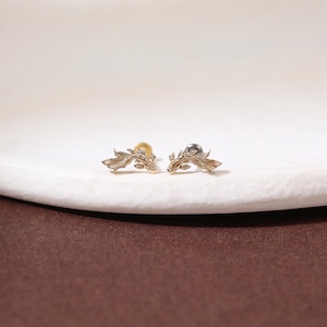 May include: A pair of gold-toned stud earrings, each shaped like a swimming fish with intricate, textured fin details. The earrings feature a curved, elongated body design with a metallic finish, showcasing fine engraved lines that mimic scales and fins.