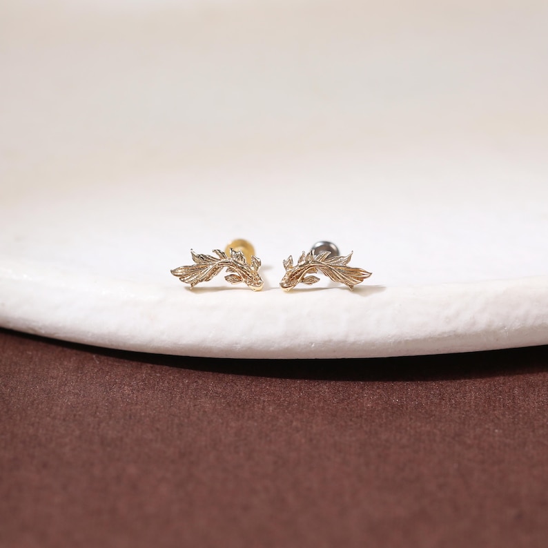 May include: A pair of gold-toned stud earrings, each shaped like a swimming fish with intricate, textured fin details. The earrings feature a curved, elongated body design with a metallic finish, showcasing fine engraved lines that mimic scales and fins.