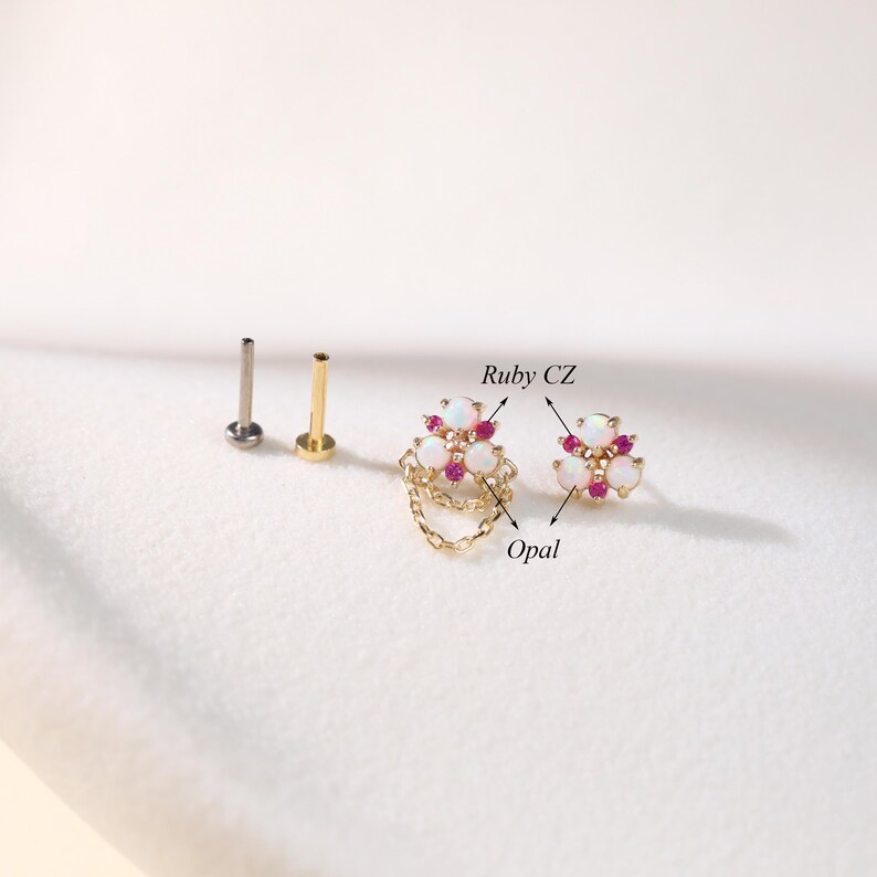 May include: A collection of gold and silver-toned body jewelry, including two studs and two decorative pieces. The decorative pieces feature opal and ruby CZ stones in a floral design, with one piece having a chain. The jewelry is set against a white background.