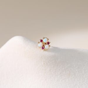 May include: A gold-colored stud earring featuring a floral design. The earring has three white opal stones and three smaller, round, red stones. The earring is set on a white surface.