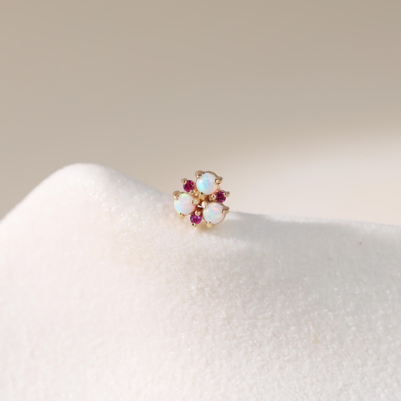 May include: A gold-colored stud earring featuring a floral design. The earring has three white opal stones and three smaller, round, red stones. The earring is set on a white surface.