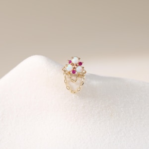 May include: A gold-colored earring with a cluster of three white opal stones and small red gems. The earring features a dangling chain design. The jewelry is set against a soft, white background.