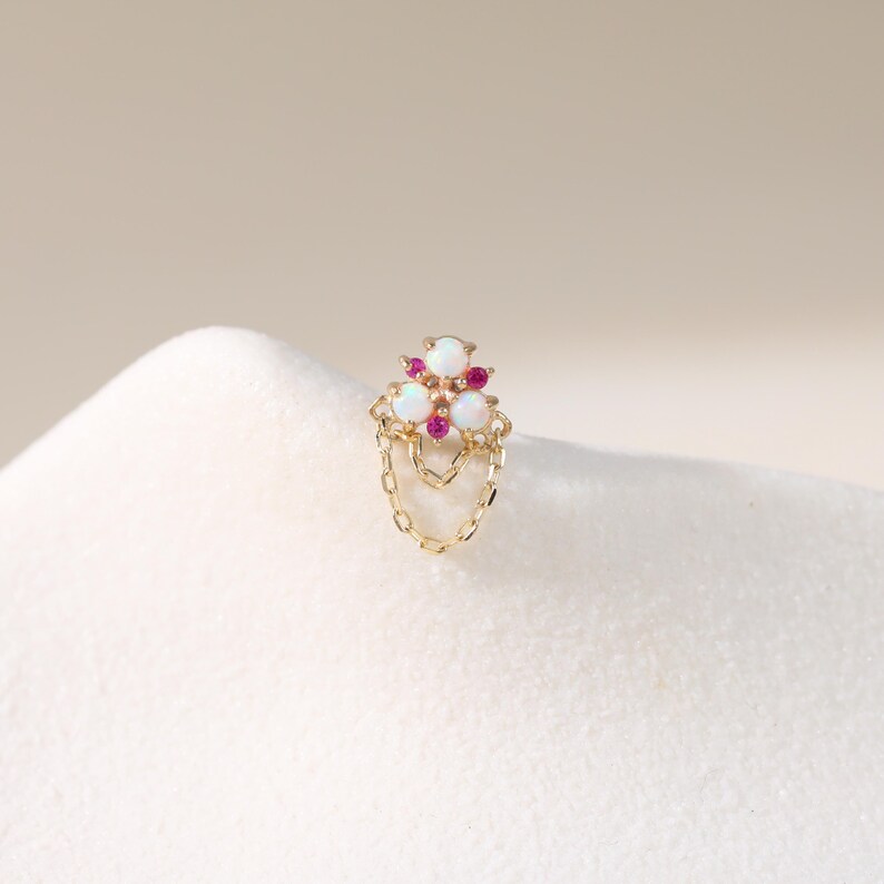 May include: A gold-colored earring with a cluster of three white opal stones and small red gems. The earring features a dangling chain design. The jewelry is set against a soft, white background.