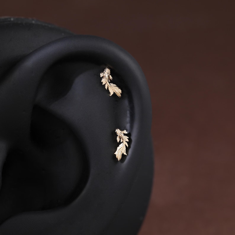 May include: Two gold-toned metal earrings shaped like koi fish, each featuring detailed fins and scales. These small, delicate stud earrings are positioned along the curve of a dark, neutral-colored ear model to display their placement.
