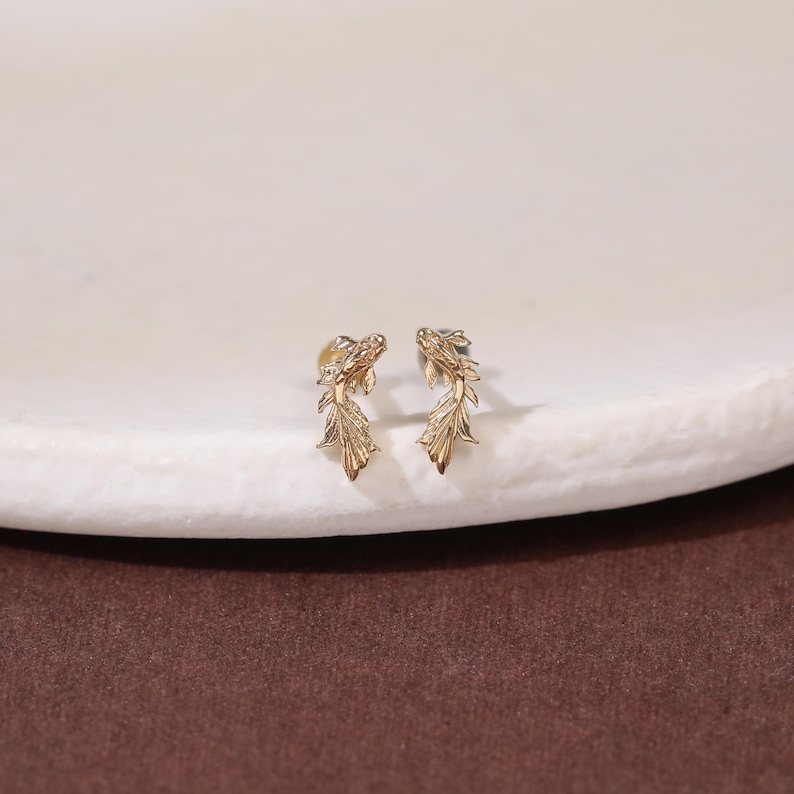 May include: A pair of gold-toned stud earrings shaped like swimming fish with detailed fins and scales. The earrings feature a metallic finish and are designed with a post-back mechanism, showcasing a slender, elongated silhouette that mimics the movement of a fish.