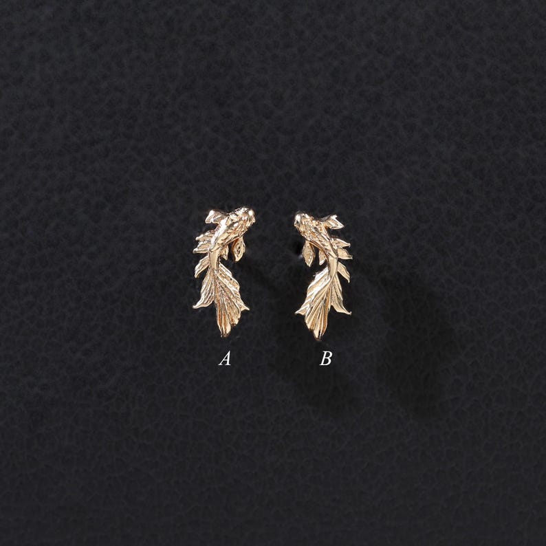 May include: Two gold-toned metal stud earrings shaped like koi fish, each featuring intricate scale and fin detailing. The left earring is labeled with the letter A, and the right earring is labeled with the letter B, both displayed clearly.