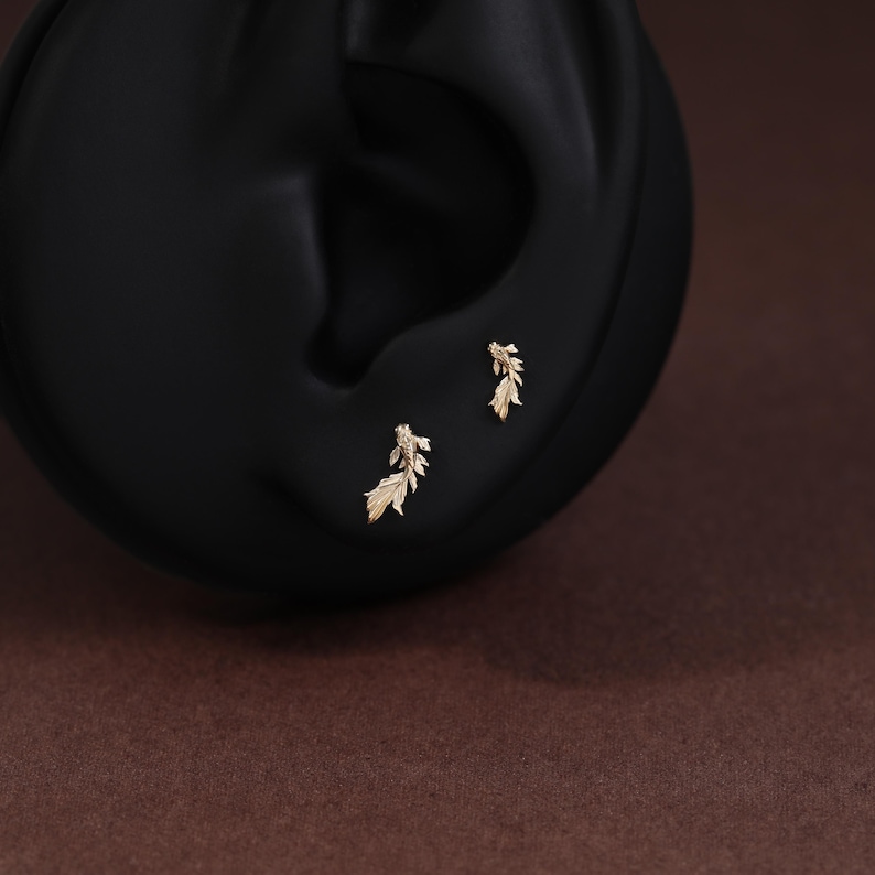 May include: A pair of small gold stud earrings shaped like koi fish, each featuring detailed fins and scales. The earrings are positioned on a black display form, showcasing the intricate metalwork and the delicate, flowing design of the fish bodies.
