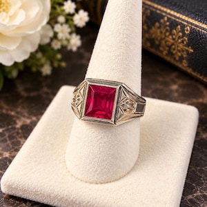 May include: A silver-toned ring featuring a large, rectangular, faceted red gemstone set in the center. The metal band displays intricate geometric engravings and milgrain detailing on the shoulders, framing the vibrant red stone securely.