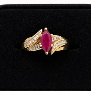 May include: A gold ring featuring a central marquise-cut deep red ruby gemstone. The band is accented with two curved rows of small, clear round-cut diamonds set into the gold, creating a bypass-style design that frames the central stone.