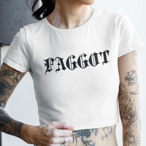 May include: White crop top with the word "FAGGOT" printed in black gothic font. The shirt has a crew neck and short sleeves. The text is centered on the front of the shirt.