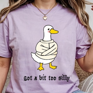 May include: A light purple t-shirt featuring a graphic of a white duck wearing a beige straightjacket with a yellow beak and feet. The duck has pink cheeks. Below the graphic, the text reads "got a bit too silly" in a black gothic font.
