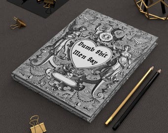 Può includere: Un taccuino rettangolare con copertina rigida caratterizzato da un'intricata illustrazione in bianco e nero in stile barocco con figure alate e volute ornamentali. Un emblema centrale a forma di cuore contiene il testo "Dumb Shit men say".