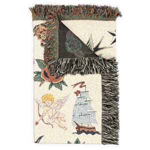 May include: Cream-colored woven throw blanket featuring classic tattoo-style illustrations. The design includes a rose, a ship, an angel with a bow and arrow, and a star. The blanket has a fringed edge with a mix of colors.