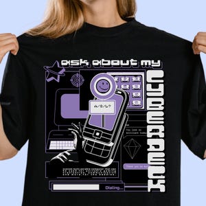 May include: A black t-shirt featuring a purple and white graphic print with the text "ASK ABOUT MY CYBERDECK". The design includes a vintage mobile phone, a computer monitor, a keypad, and a chat bubble with the text "A/S/L?" and "Dialing..."