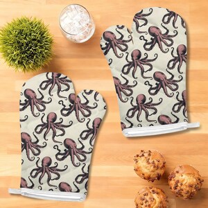 May include: Two oven mitts with a cream-colored background and a repeating pattern of purple octopuses. The mitts are quilted and have a white loop for hanging. The image also shows a small green plant, a glass of water, and some muffins.