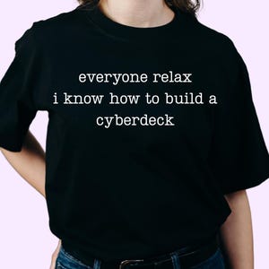 May include: A black short-sleeved t-shirt featuring white text printed on the chest that reads, everyone relax i know how to build a cyberdeck. The garment has a crew neckline and a standard fit, constructed from a solid black fabric material.