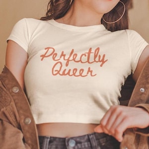 Peut inclure: Haut court crème avec l'inscription "Perfectly Queer" en script rouge-orangé. Le t-shirt a des manches courtes et un col rond. Le modèle porte une veste marron et un jean foncé.