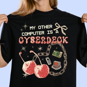 May include: A black t-shirt featuring the text "MY OTHER COMPUTER IS A CYBERDECK" in white and red bubble letters. The graphic includes two red cherries, a beige chain, a small circuit board, a vintage computer, and a mobile phone.