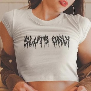 May include: White crop top with the text "SLUTS ONLY" in a black, gothic font. The shirt has short sleeves and a crew neck. The model is wearing a brown jacket.