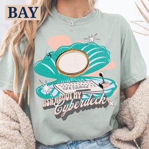 Könnte beinhalten: Hellgrünes T-Shirt mit Retro-Grafik. Das Design zeigt eine Tastatur, einen Joystick und den Text "ASK ABOUT MY Cyberdeck" in einer Vintage-Schriftart. Die Grafik enthält ein goldgerahmtes Oval und Sternausbrüche.