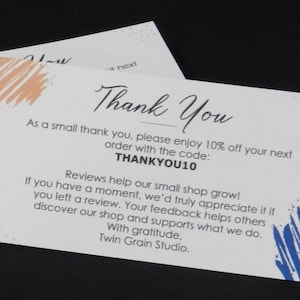 May include: A stack of white thank you cards with the text "Thank You" in elegant script. The cards have orange and blue brushstroke accents and include a discount code. The cards are from Twin Grain Studio.