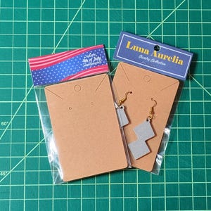 May include: Two pairs of earrings on brown cardboard earring cards, packaged in clear plastic. One card has a red, white, and blue banner that says "Happy 4th of July Independence". The other card has a blue banner that says "Luna Aurelia Jewelry Collection".