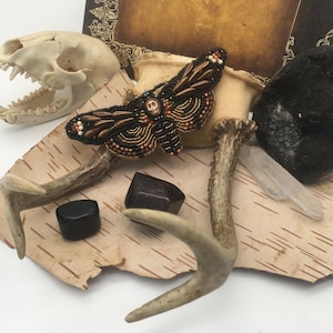 Puede incluir: Un broche de polilla de cuentas con un diseño de calavera, exhibido sobre un fondo de corteza de abedul con astas, una piedra negra y un cristal de cuarzo transparente.