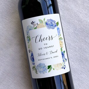 Op de afbeelding: Een wit wijnfleslabel met een blauw en wit bloemenontwerp. Het label zegt "Cheers to 20 Years! Alexis & David September 5, 202X".