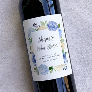 Op de afbeelding: Een wit wijnfleslabel met een blauw en wit bloemenontwerp. De tekst op het label luidt "Megan's bridal shower" en "Thank you for celebrating with us! August 22, 20XX".