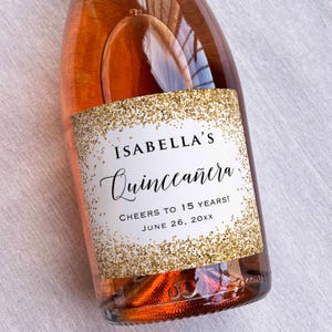 May include: A white wine bottle label with gold glitter and the text "Isabella's Quinceañera Cheers to 15 Years! June 26, 20XX"