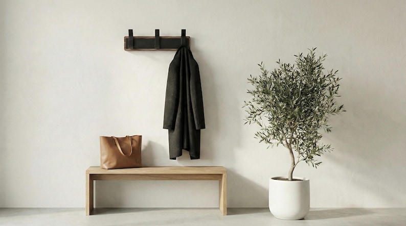 Pu&ograve; includere: Una scena d'ingresso minimalista con una panca in legno, un attaccapanni con un cappotto scuro e una borsa tote in pelle. Un albero di ulivo in vaso aggiunge un tocco di verde. Il muro &egrave; dipinto di un colore neutro.