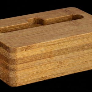 May include: A rectangular bamboo phone stand with a slot for holding a phone. The stand is made of layered bamboo and has a natural wood grain finish. The stand is designed to hold a phone upright.