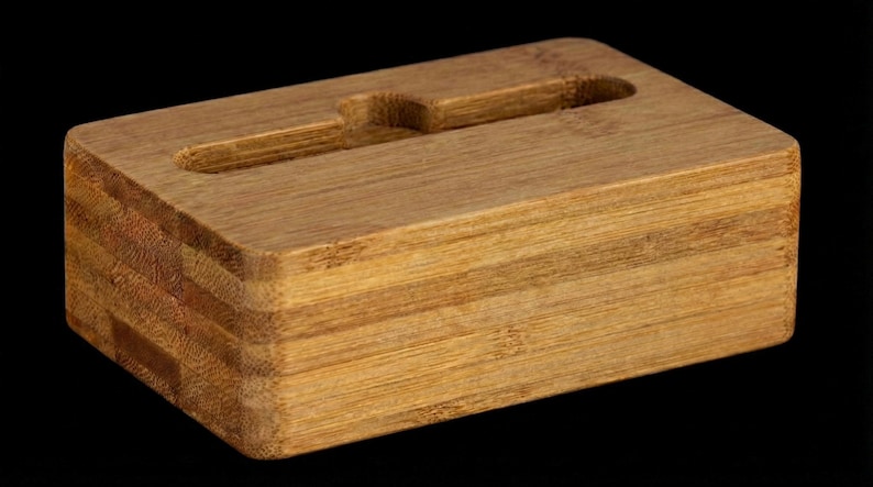 May include: A rectangular bamboo phone stand with a slot for holding a phone. The stand is made of layered bamboo and has a natural wood grain finish. The stand is designed to hold a phone upright.