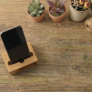 May include: A wooden phone stand holds a black smartphone. The stand is made of light-colored wood and has a slot to hold the phone upright. Small potted plants and twine are in the background, on a wooden surface.