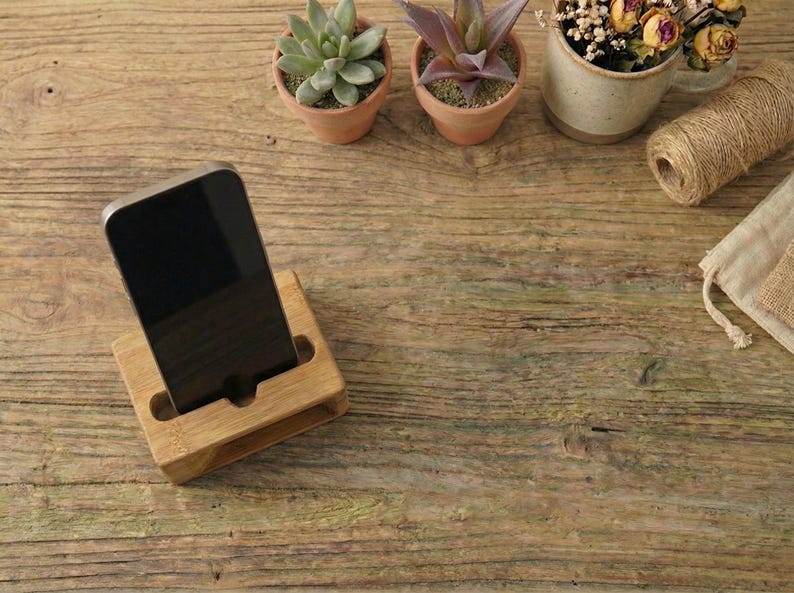 May include: A wooden phone stand holds a black smartphone. The stand is made of light-colored wood and has a slot to hold the phone upright. Small potted plants and twine are in the background, on a wooden surface.