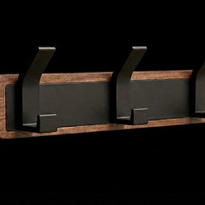 Pu&ograve; includere: Un attaccapanni da parete con tre ganci in metallo nero. I ganci sono montati su una tavola rettangolare in legno con finitura marrone scuro e un pannello centrale nero. Design minimalista e moderno.