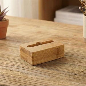 May include: A rectangular bamboo tissue box with a slot on top sits on a wooden table. Two small potted succulents and a mug with dried flowers are also on the table. A roll of twine and a small drawstring bag are also visible.
