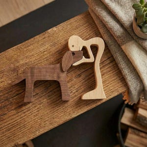 May include: Wooden dog and figure sculptures. The dog is brown, and the figure is light-colored. The figure is embracing the dog. The sculptures are on a wooden surface. A blanket and a small potted plant are in the background.